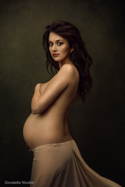 Pregnant woman in profile pose against dark background with draped fabric.
