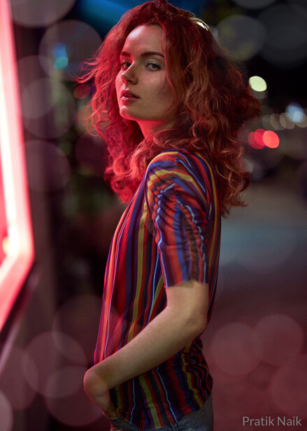 Woman with wavy red hair looking over her shoulder in neon-lit urban setting.