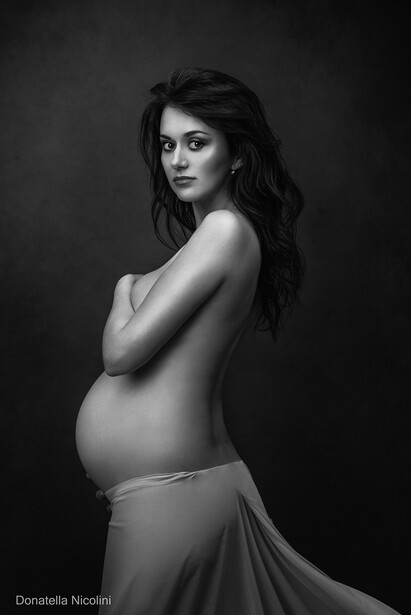 Pregnant woman in profile against dark background, wearing sheer fabric draped low.