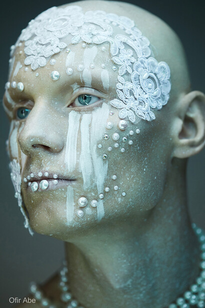 Portrait of a person with theatrical white face paint and pearl embellishments, shot in profile against neutral background.