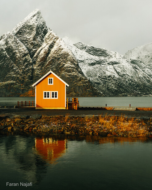 Bright yellow cabin reflected in still water with snow-capped mountain backdrop.