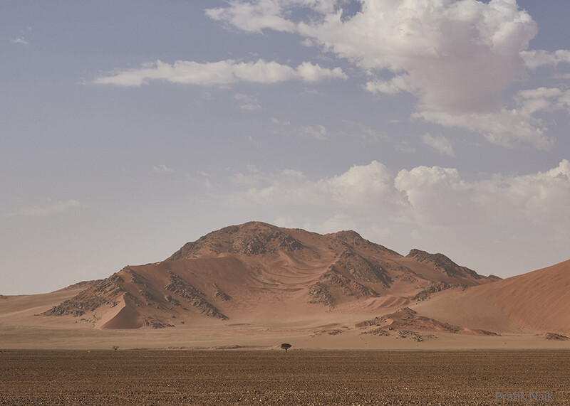 Desert landscape with isolated mountain under partly cloudy sky and sparse vegetation in foreground.