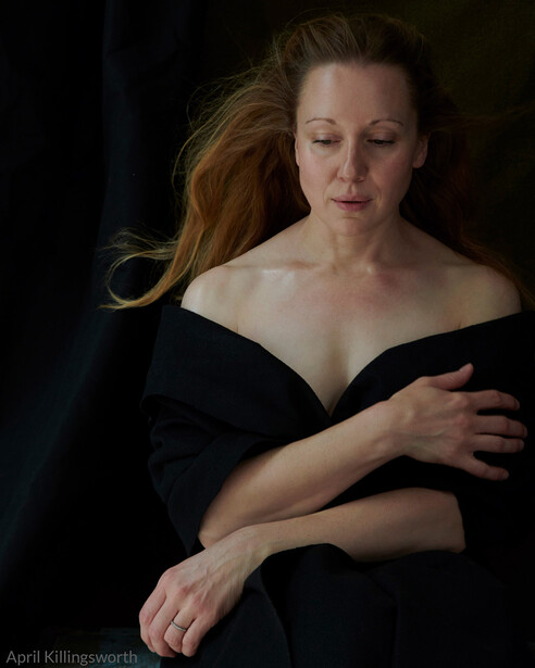 Woman in black off-shoulder top against dark background, gazing downward with composed expression.