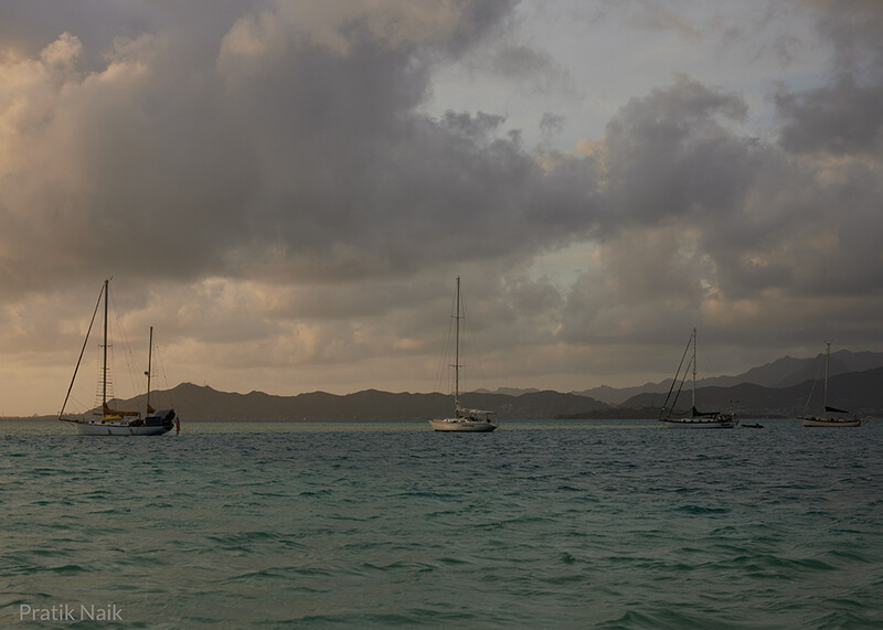 Four sailboats anchored in turquoise water with mountains in the distance under dramatic cloudy sky.