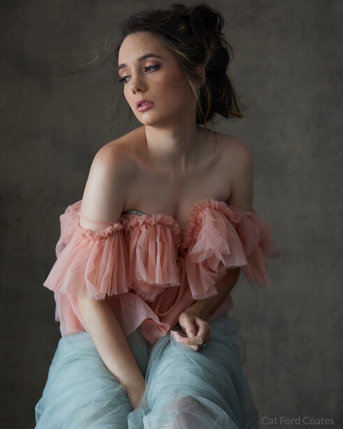 Woman in pink ruffled off-shoulder top and light blue jeans against neutral gray backdrop.