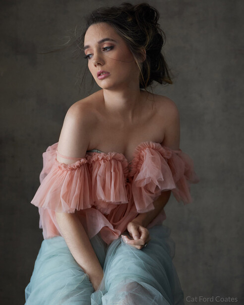 Young woman in ruffled off-shoulder pink top and light blue jeans gazes to the side against neutral gray background.