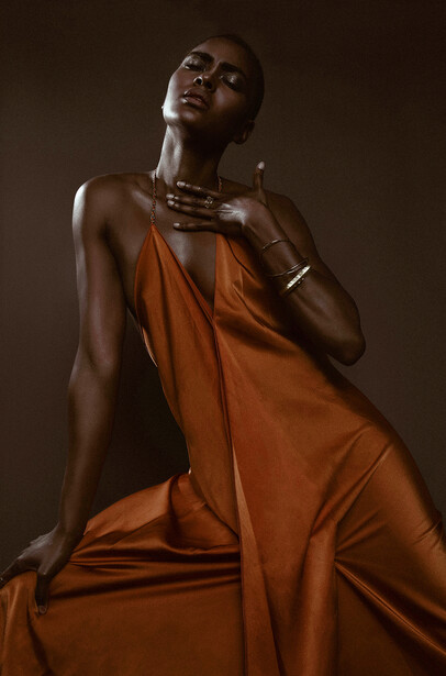 Woman in rust-colored draped fabric against warm brown background, looking upward with contemplative expression.