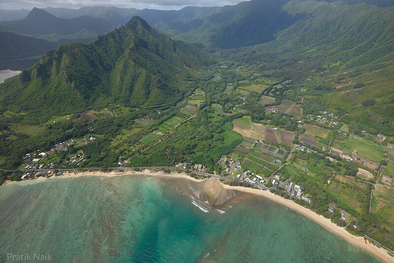 Aerial view of a coastal valley with lush green mountains, taro fields, and a curved sandy beach with turquoise waters.