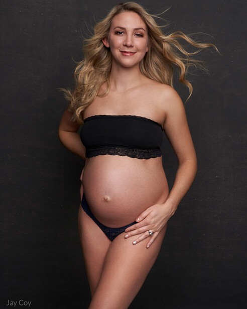 Pregnant woman in studio portrait wearing black bandeau top against dark background.