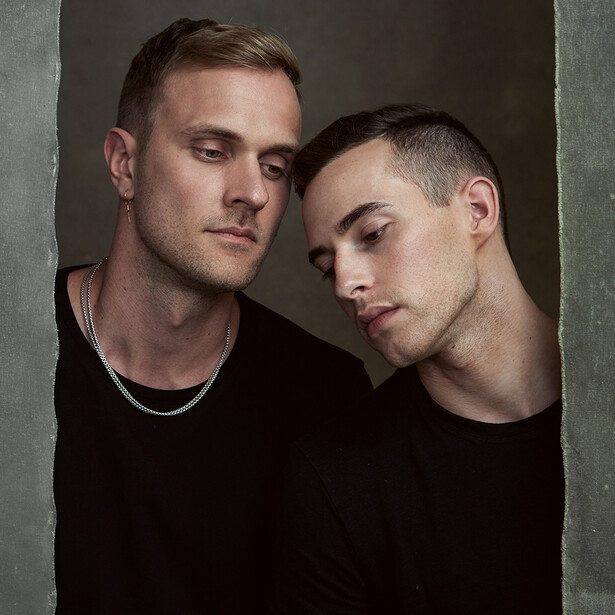 Two men in black shirts posed closely together against a neutral gray backdrop.