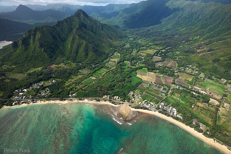 Aerial view of a lush Hawaiian valley with turquoise waters, sandy beach, green mountains, and scattered settlements.