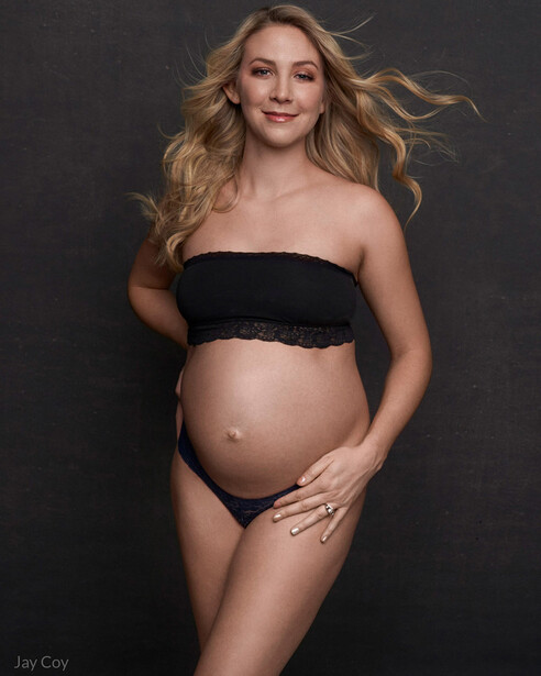 Pregnant woman in studio portrait wearing black bandeau top against dark background.