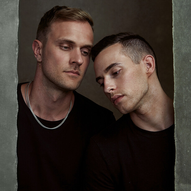 Two men in black shirts posed close together against a neutral backdrop.