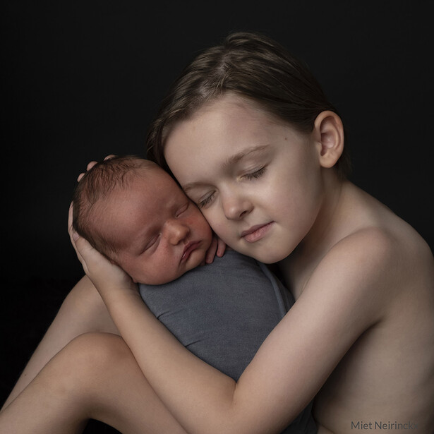 Older child holding and nuzzling a sleeping newborn against shoulder in intimate studio portrait.