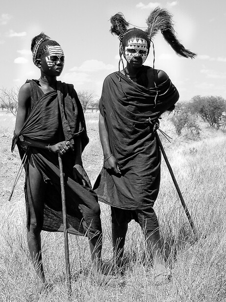 Two people in traditional African dress and beaded headdresses standing in grassland, photographed in black and white.