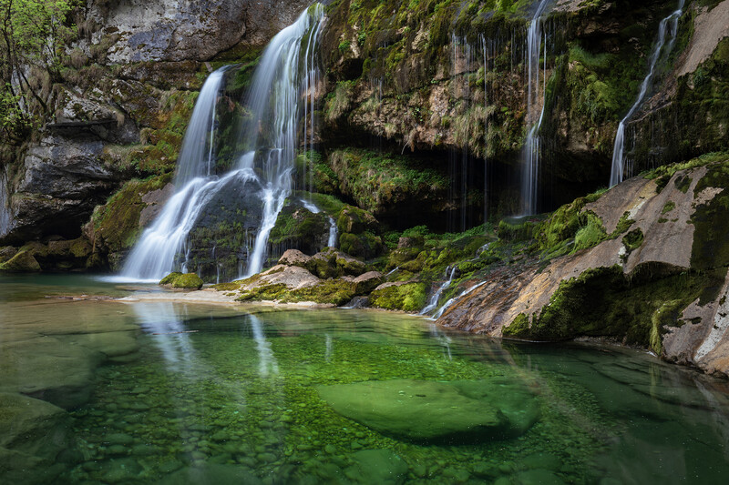 Cascading waterfalls flow into an emerald pool surrounded by moss-covered rocks and lush vegetation.