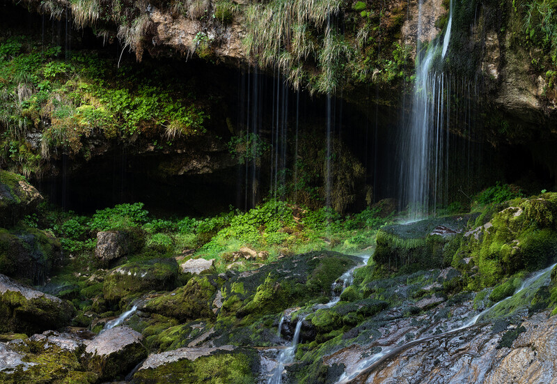 Waterfall cascading over moss-covered rocks in a lush forest canyon with dense green vegetation.