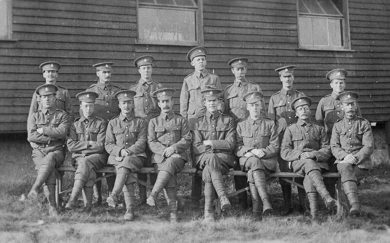 Formal group portrait of uniformed soldiers arranged in three rows against a wooden building.