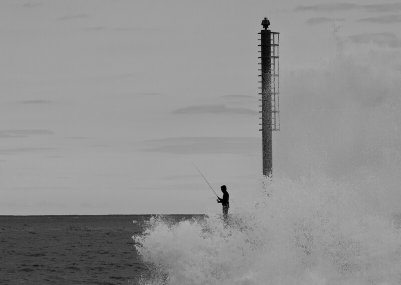 Surfer riding a large wave near a tall pier structure in black and white.