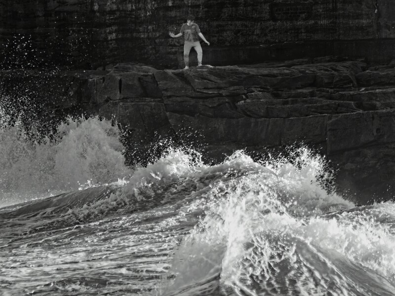 Silhouetted figure standing on a rocky outcrop overlooking turbulent ocean waves in black and white.