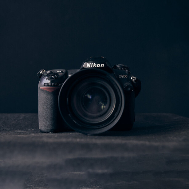 Nikon D500 DSLR camera with attached lens on dark textured surface against dark teal background.