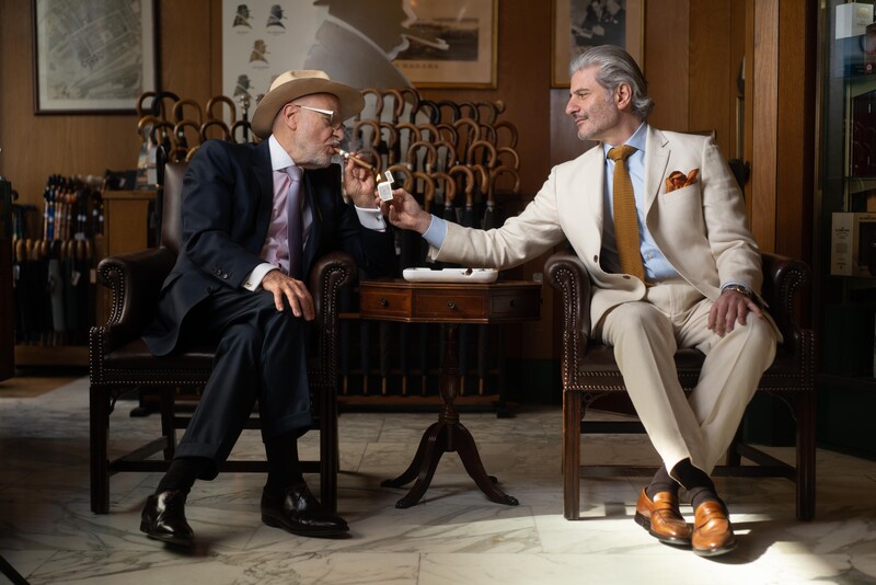 Two men in suits conversing in a wood-paneled interior, seated in leather chairs.