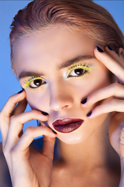 Beauty portrait with bold yellow eyeshadow and burgundy lips against blue background.
