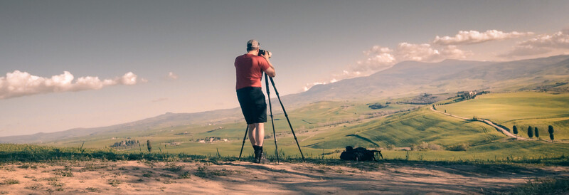 Photographer on tripod in rolling landscape hills during golden hour with warm sunlight and clouds.