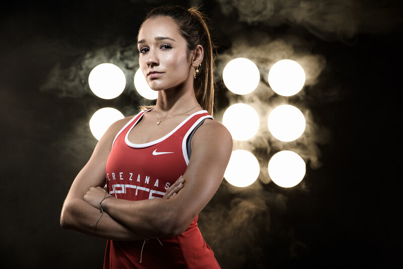 Female athlete in red tank top with arms crossed, standing against blurred stage lights.