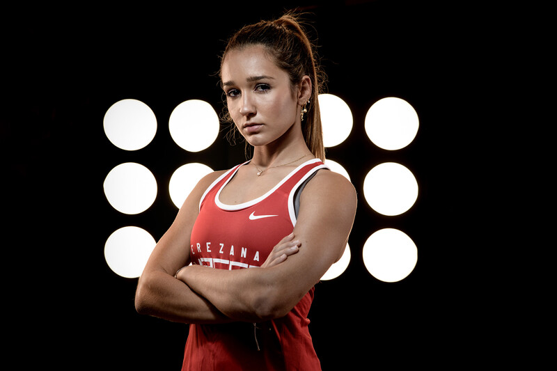 Female athlete in red tank top with arms crossed, positioned against black background with ring lights.