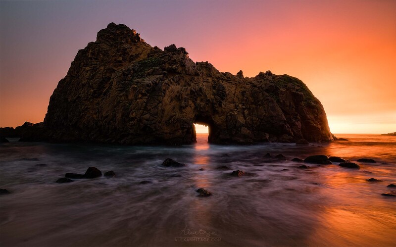 Large rocky sea arch silhouetted against a golden sunset with waves washing across the beach.