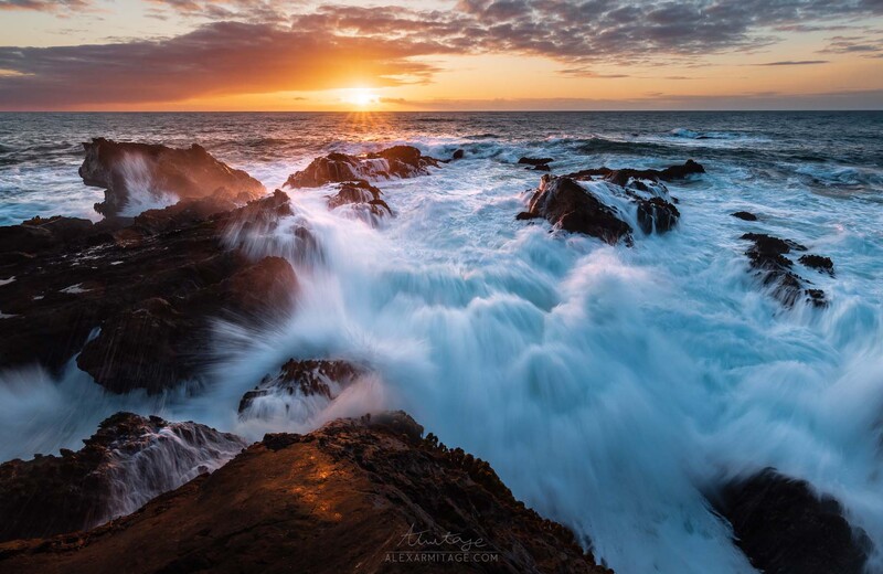 Rocky coastline with crashing waves at golden hour sunset, waves creating white spray over dark boulders.