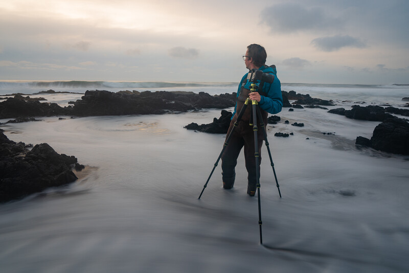 Photographer with tripod standing in shallow water among coastal rocks during golden hour.
