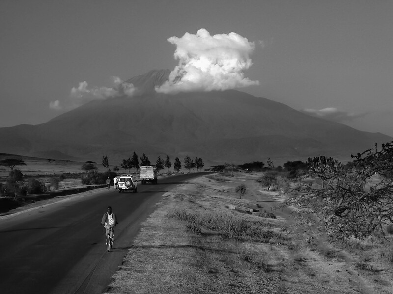 A cyclist rides on a paved road toward a volcano shrouded in clouds and smoke, with a truck ahead and sparse vegetation on either side.
