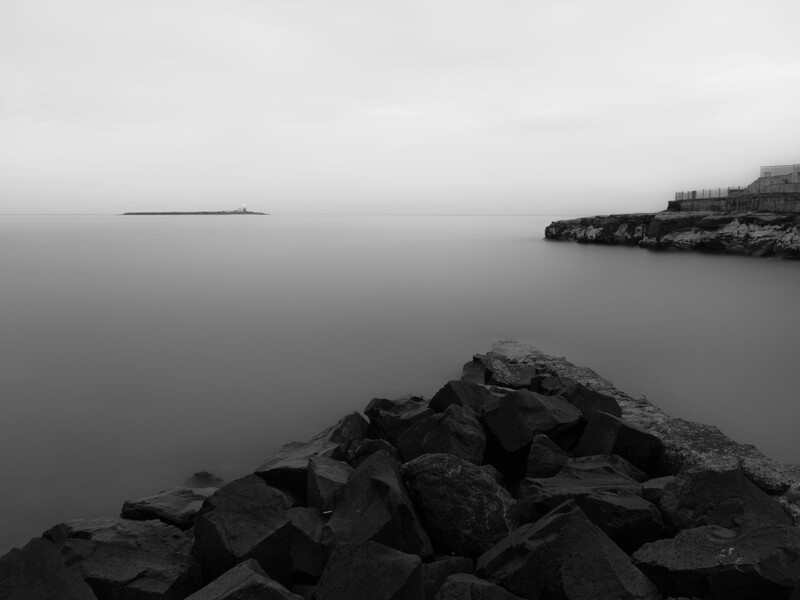 Rocky jetty in foreground with calm, mirror-like water and distant shoreline structures under overcast sky.