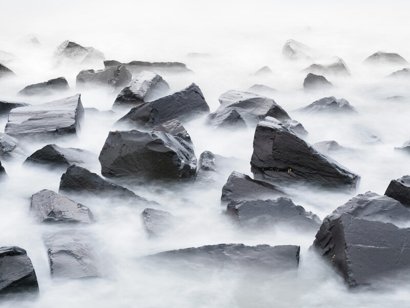 Dark rocks emerging from smooth, flowing white water in a long-exposure seascape.