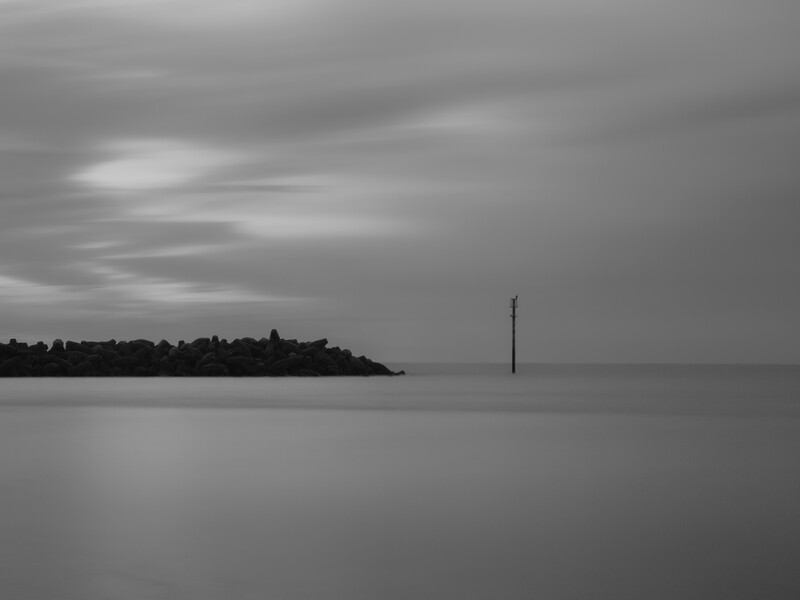 Minimalist seascape with a solitary wooden post rising from calm water beneath an overcast sky.