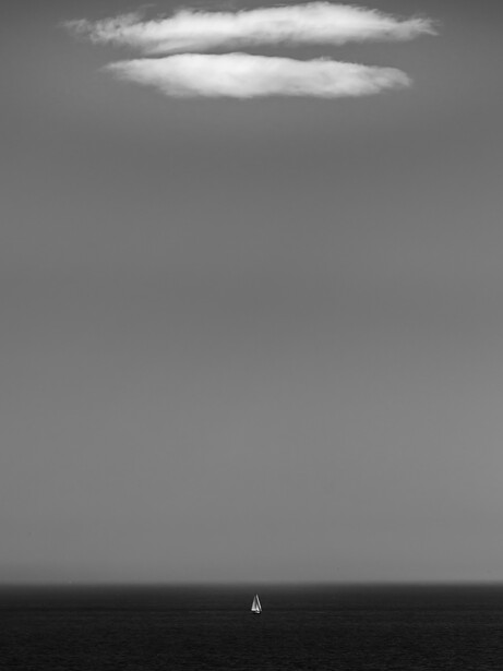 Minimalist black and white seascape with a single sailboat on the horizon beneath an overcast sky.