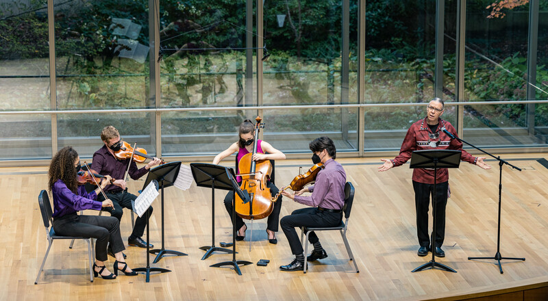 String quartet performing in a modern venue with large windows overlooking trees.