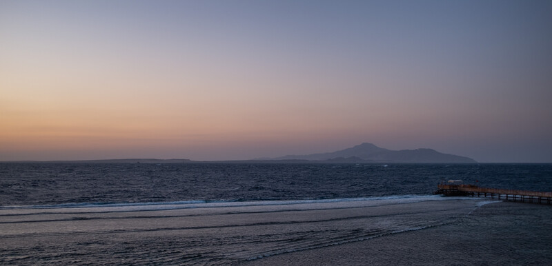 Volcanic mountain silhouetted against twilight sky above a darkening seascape with pier.