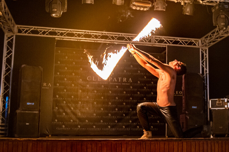 Fire performer on stage spins flaming prop overhead during nighttime performance under theatrical lighting.