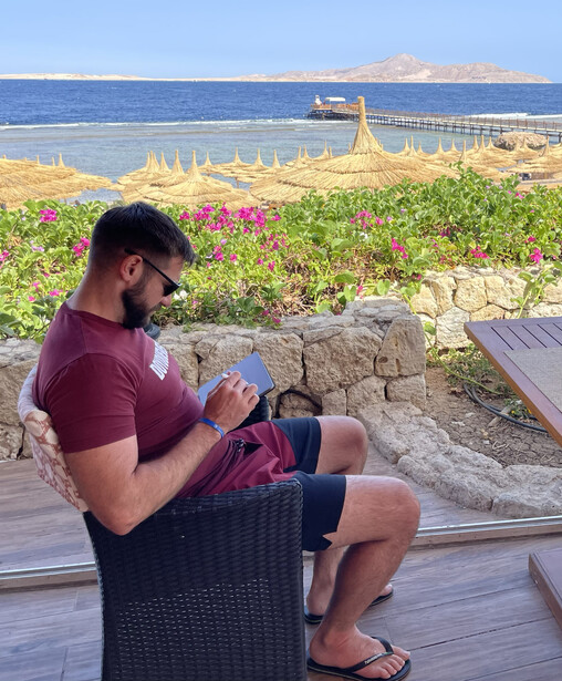 Man in maroon shirt sitting outdoors with coastal landscape and flowering plants in background.