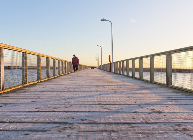 Person jogging on a wooden boardwalk at sunset with metal railings and street lamps lining both sides.