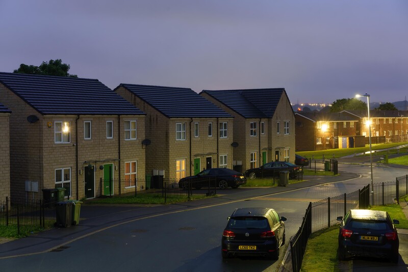 Residential street at dusk with stone terraced houses illuminated by exterior lights and street lamps.