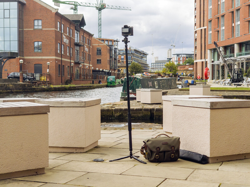 Street photography setup with tripod-mounted camera on urban waterfront plaza surrounded by brick buildings.