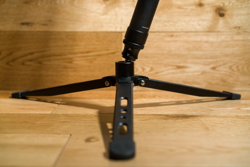 Black microphone on tripod stand with extended legs, positioned on wooden floor.