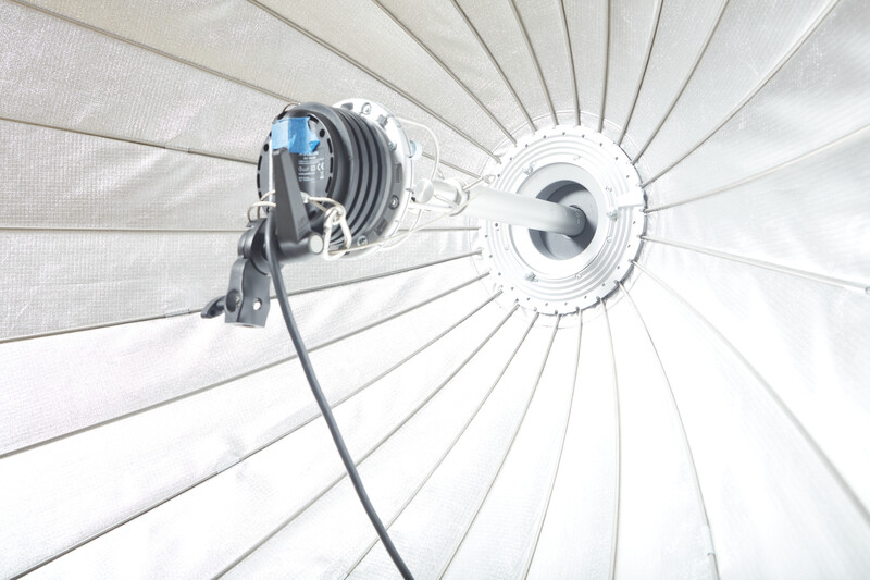 Studio strobe light with reflector and diffusion umbrella mounted overhead against white background.