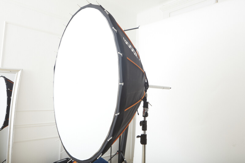 Large circular softbox with diffusion panel mounted on a studio light stand against white wall.