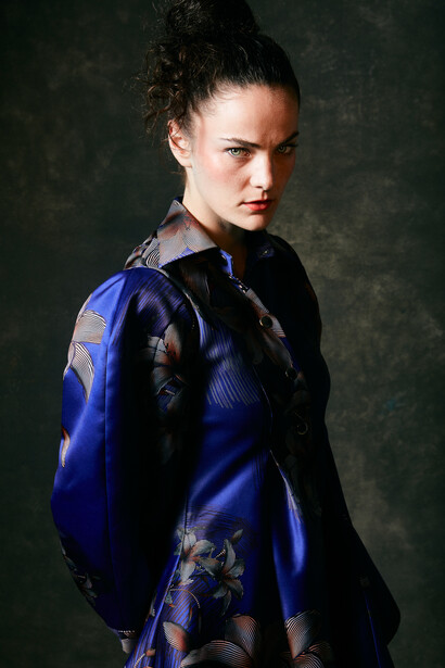 Woman in blue embroidered jacket looking over shoulder against dark background.