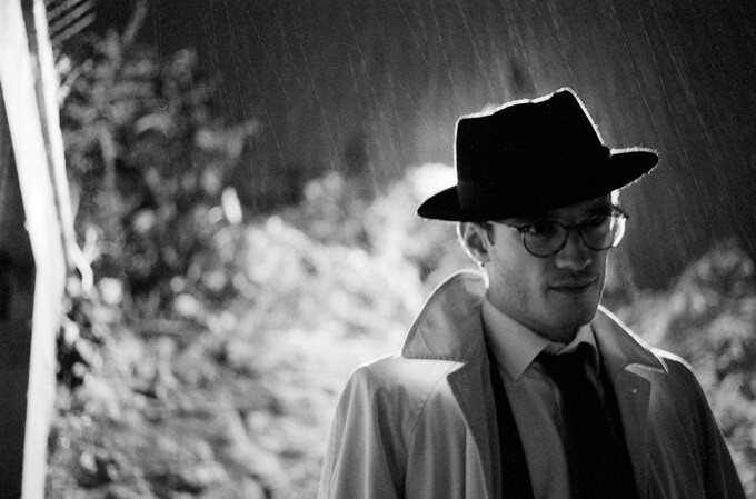Man in fedora and glasses wearing a light jacket and dark tie, photographed in profile against a rainy background.
